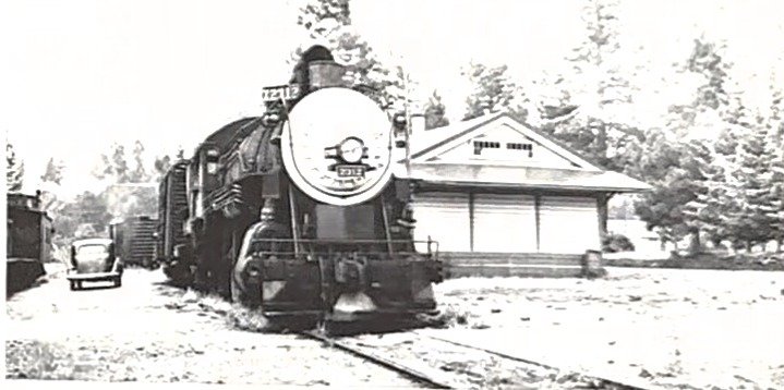 An old black and white photograph of a steam locomotive in front of a small station building, cut from a page and squared up
